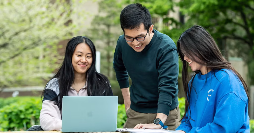 Three students sitting at a picnic bench outside and looking at a laptop screen.