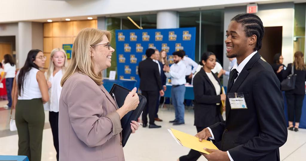 A male business student wearing professional clothing engages in conversation with a female recruiter.